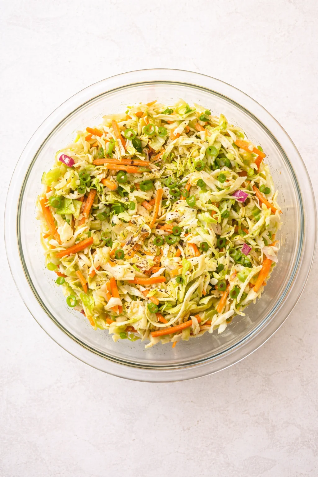 Top-down view of a glass bowl filled with shredded cabbage, carrots, peas, red onion, and green onions.