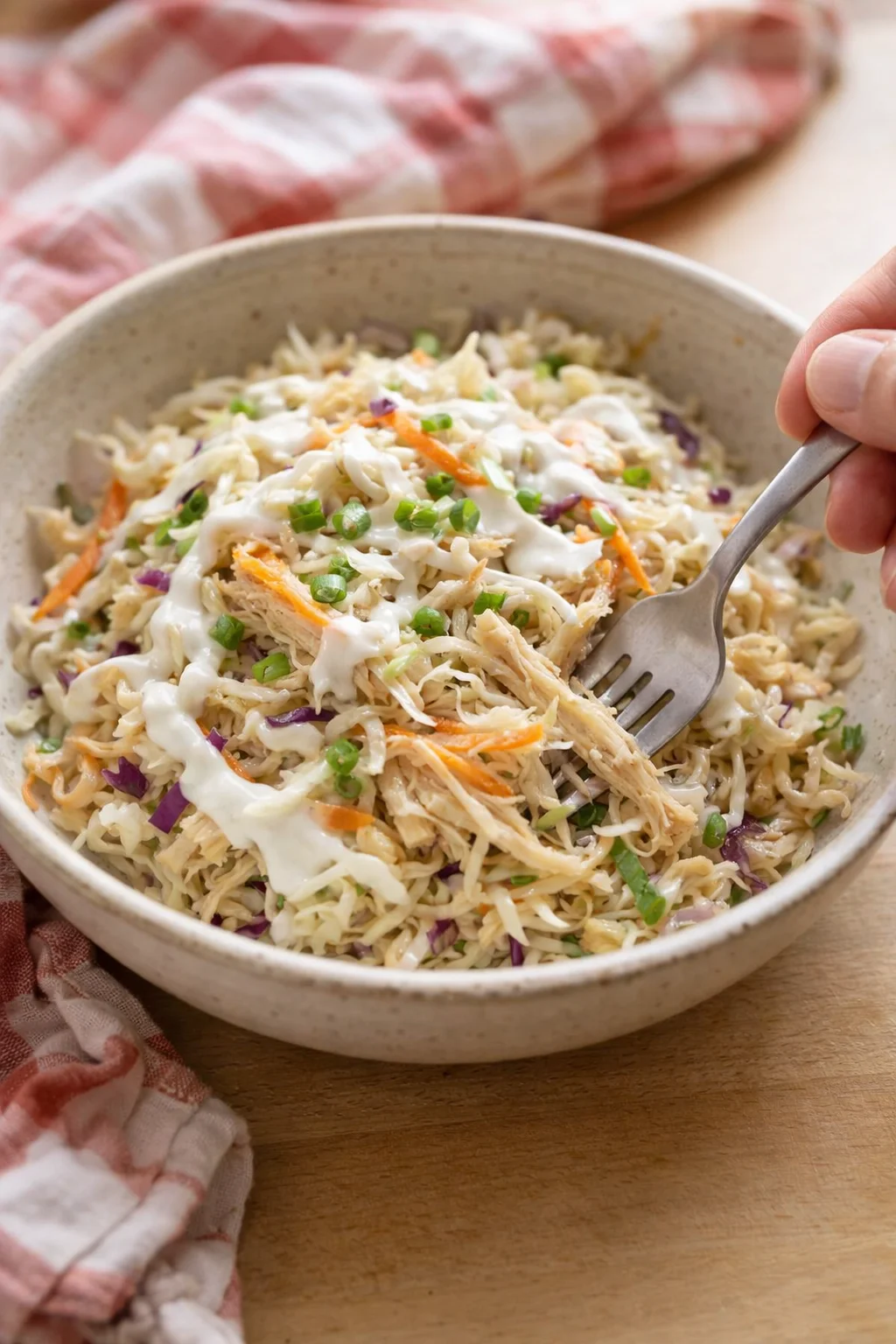 A bowl of creamy shredded chicken salad with cabbage, carrots, and green onions, being scooped with a fork.