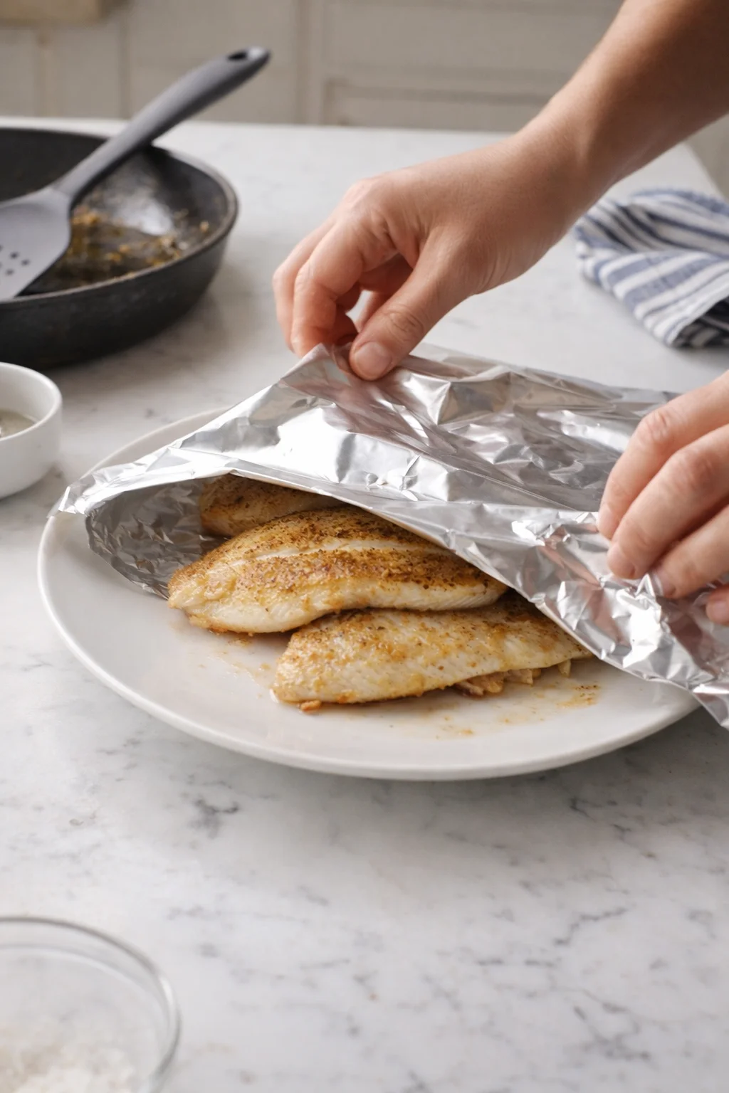 Hands peel back foil to reveal seasoned fish fillets on a white plate.