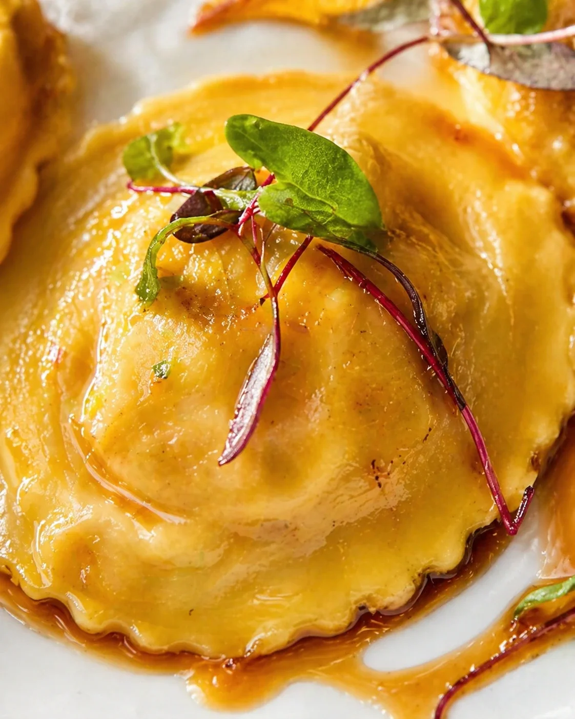 Close-up of a glossy golden pastry on a white plate, topped with green and purple microgreens.