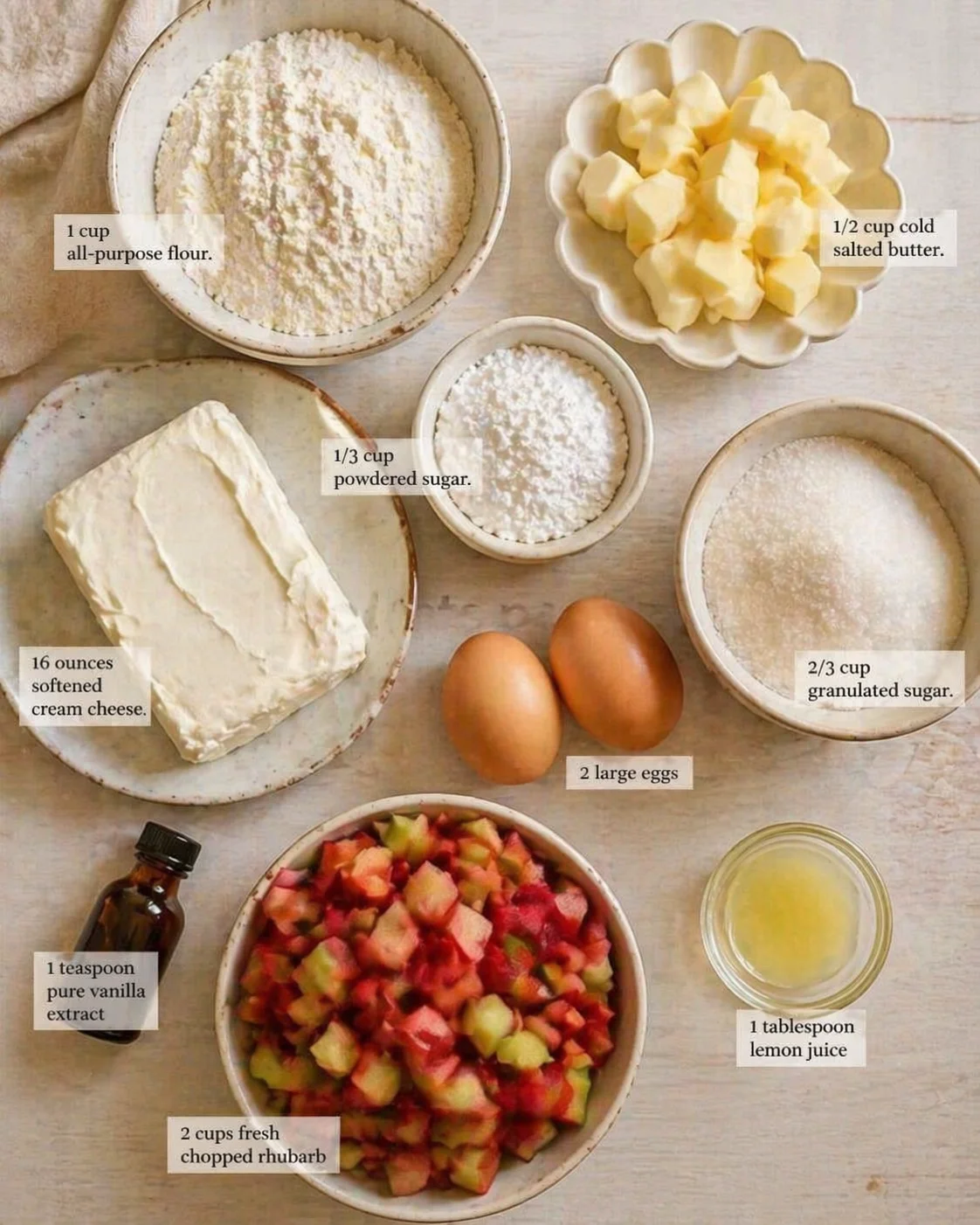 Overhead view of baking ingredients arranged on a light wood surface: flour, butter, powdered sugar, cream cheese, rhubarb, eggs.