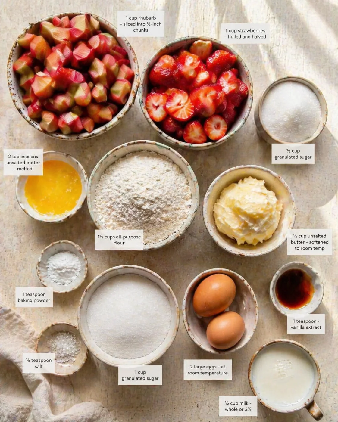 Overhead view of speckled bowls with rhubarb, strawberries, flour, butter, eggs, and sugar