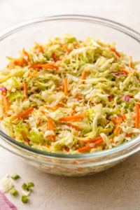 Glass bowl of shredded cabbage and carrot slaw with green onions on a beige surface.