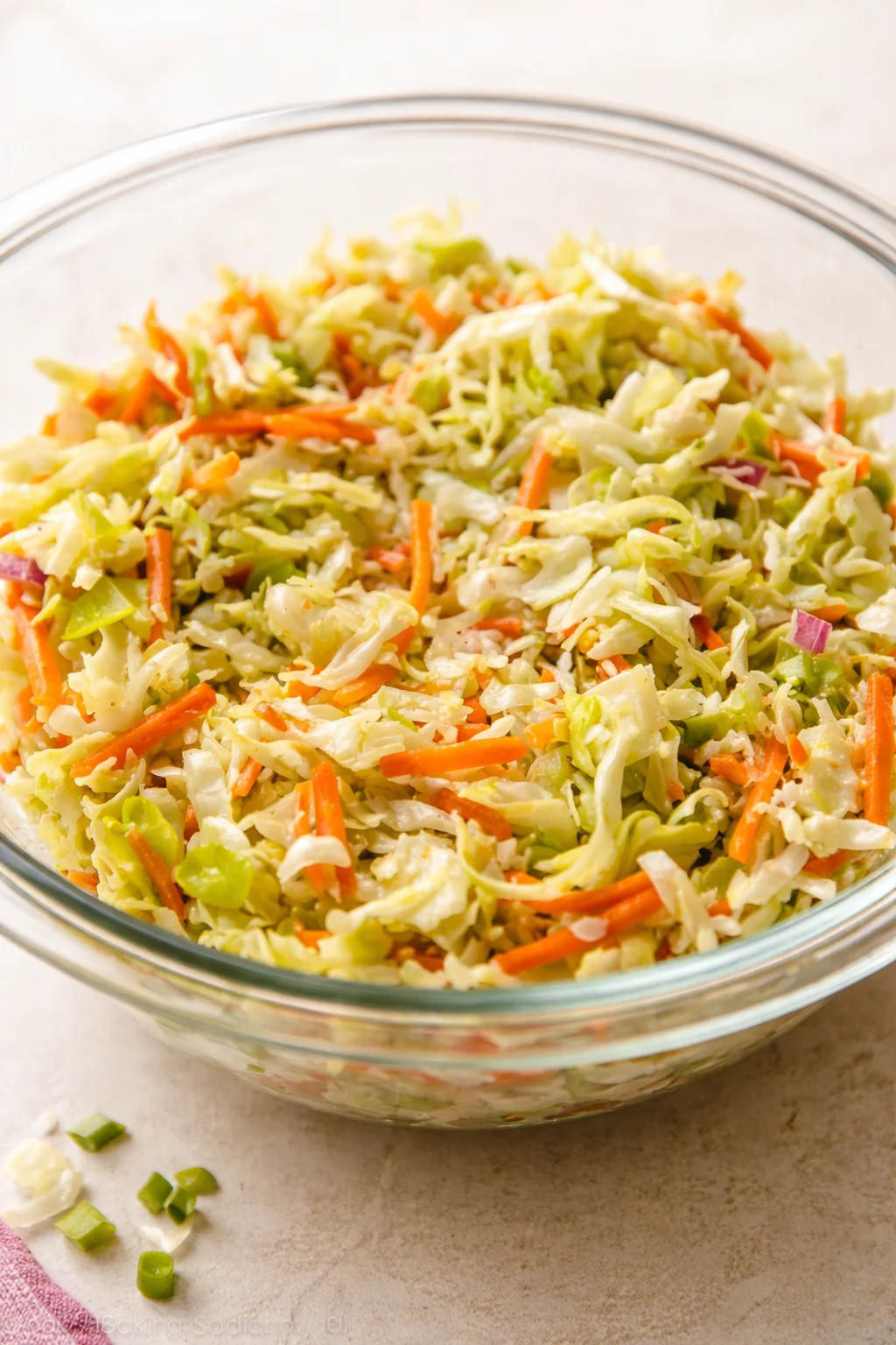 Glass bowl of shredded cabbage and carrot slaw with green onions on a beige surface.