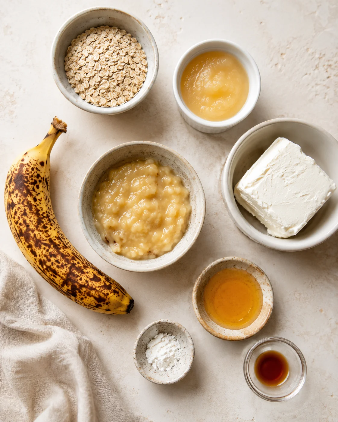 Top-down view of a flat lay with banana, oats, creamy mixture, cream cheese, and small bowls on a beige countertop.