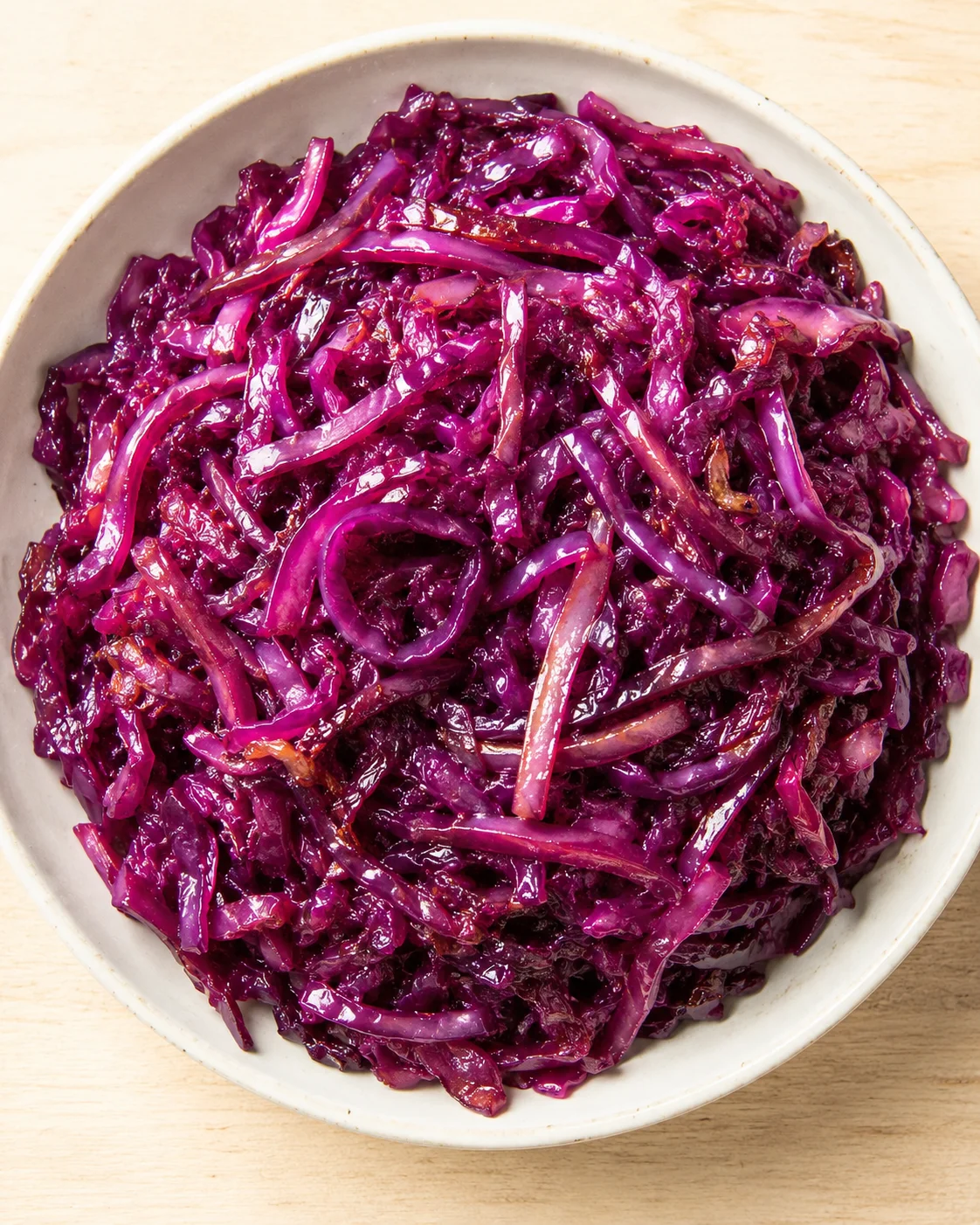 A white bowl of glossy braised red cabbage ribbons on a light wooden surface.