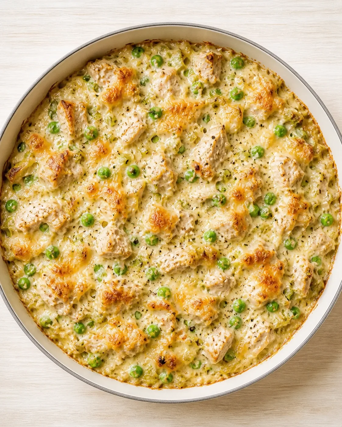round pan of creamy chicken and peas casserole with golden-brown top on a light wooden surface