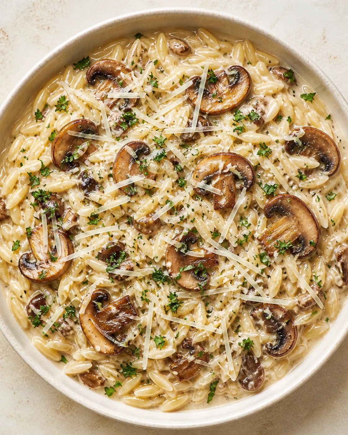 Creamy mushroom-orzo pasta in a white bowl topped with parmesan shavings and parsley