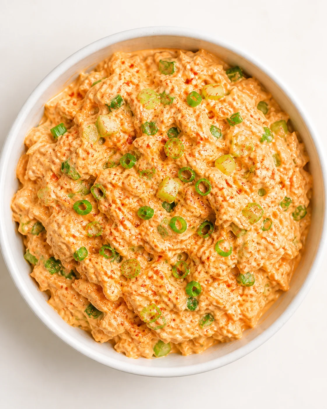 orange creamy dip in a white bowl topped with green onion rings and red chili flakes