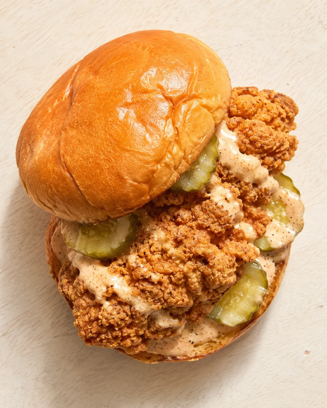 Golden brioche bun sandwich with crispy fried chicken, pickles, and creamy sauce on a light wooden surface