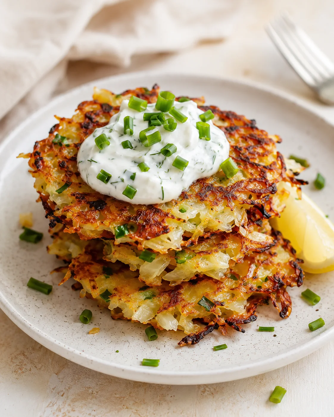 Stack of crispy potato latkes topped with dill-sour-cream and chopped chives on a beige plate.