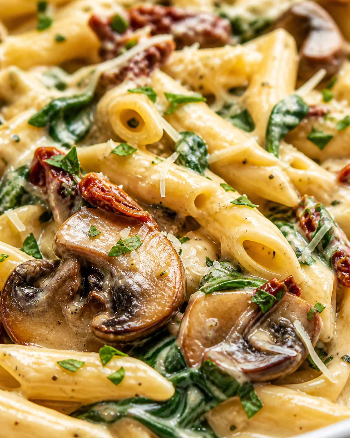 close-up of penne pasta with cream sauce, mushrooms, spinach, and sun-dried tomatoes, garnished with parsley