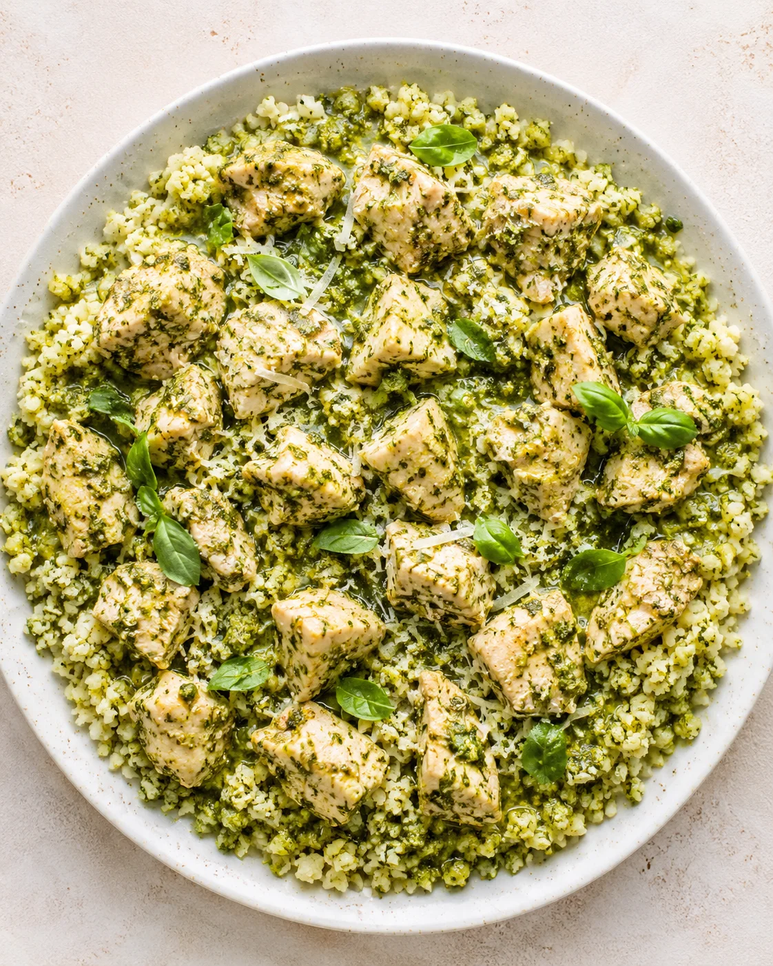 top-down view of a round plate with herb-crusted chicken cubes on green pesto couscous, garnished with basil
