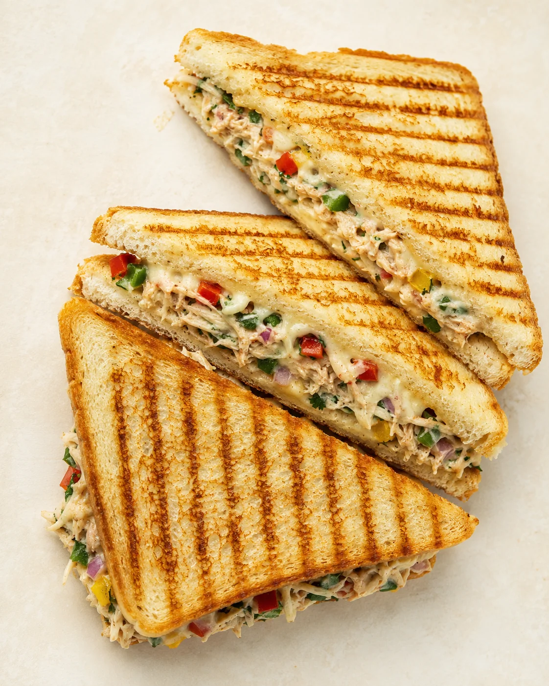 three triangular grilled tuna salad sandwiches with colorful peppers on a pale background