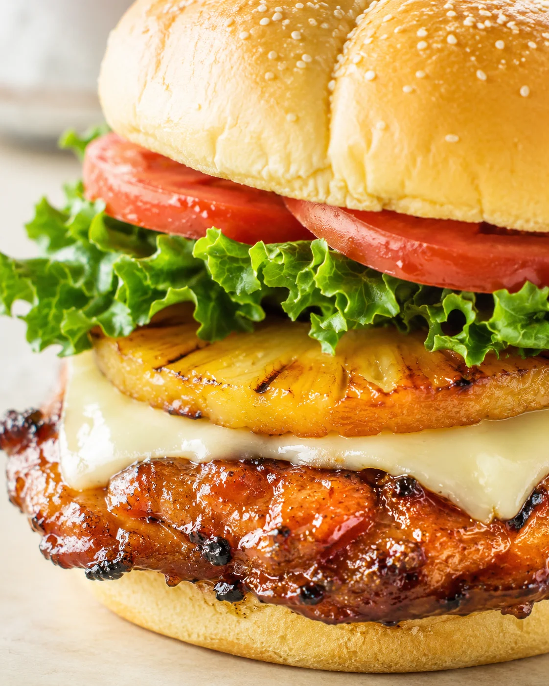 Close-up of a stacked Hawaiian-style burger with sesame bun, tomato, lettuce, grilled pineapple, cheese, and a juicy patty.