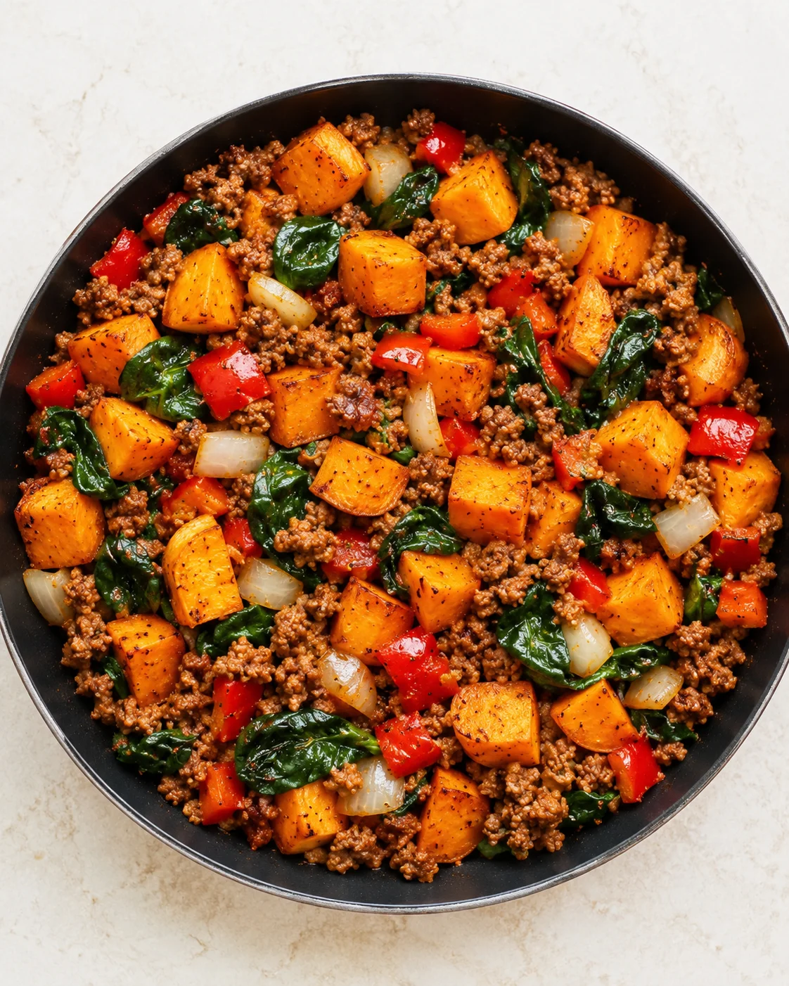 top-down view of a skillet filled with browned ground beef, cubed orange sweet potatoes, onions, red peppers, and wilted spinach.