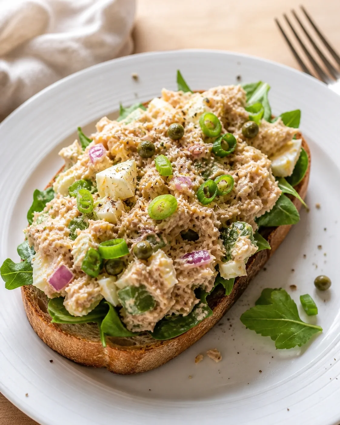 open-faced toast topped with creamy tuna salad, greens, and scallion slices on a white plate
