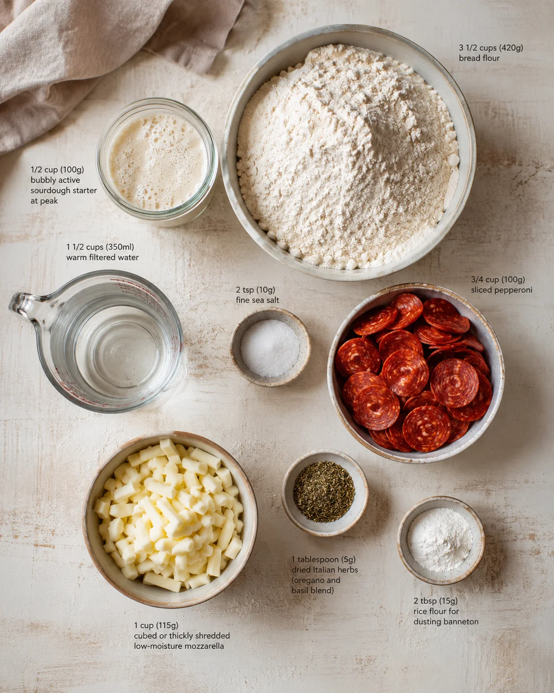 overhead view of pizza ingredients arranged on a light countertop including flour, water, starter, mozzarella, pepperoni, and herbs