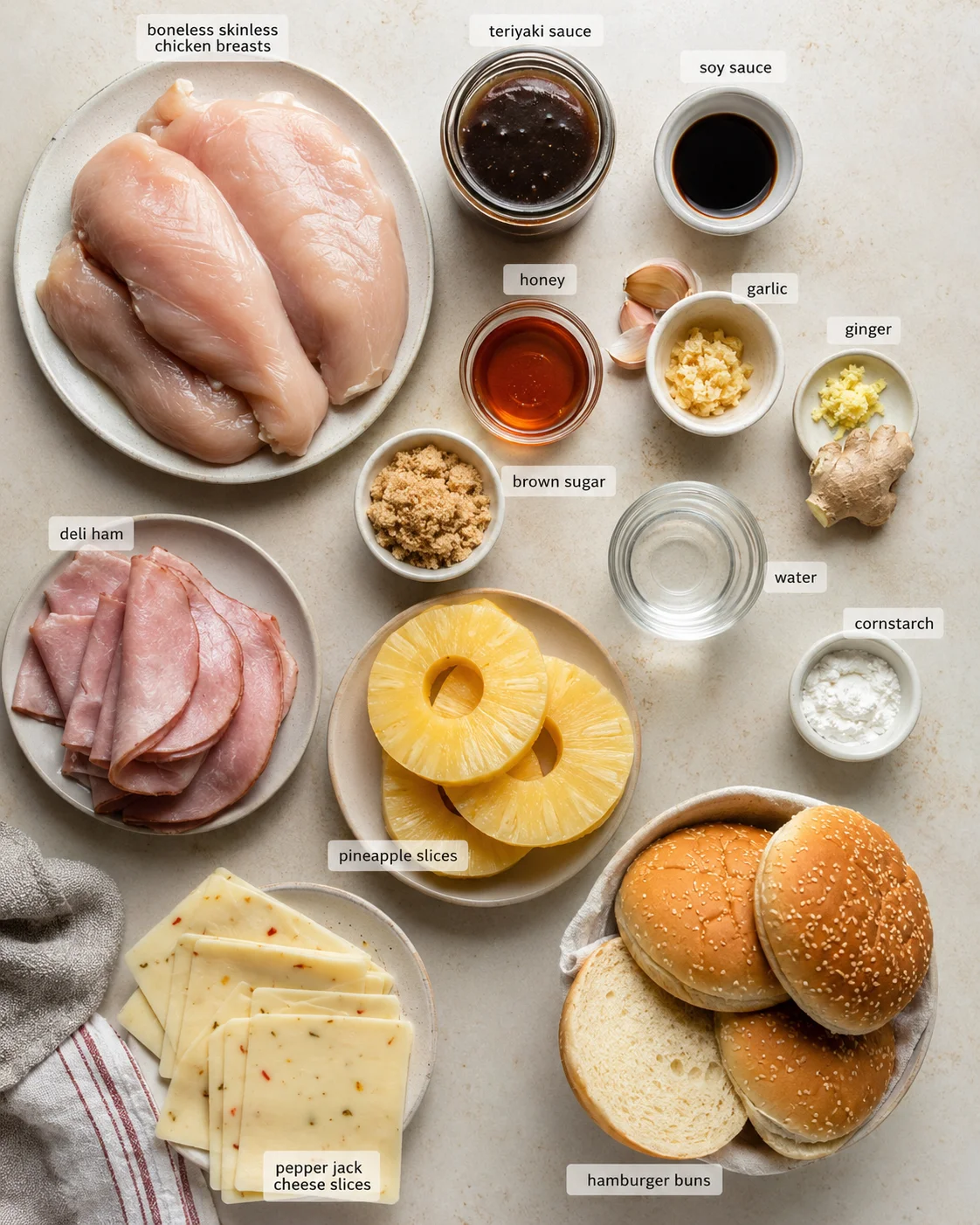 Top-down view of chicken breasts, pineapple slices, ham, cheese, buns, and labeled sauces arranged for a recipe.