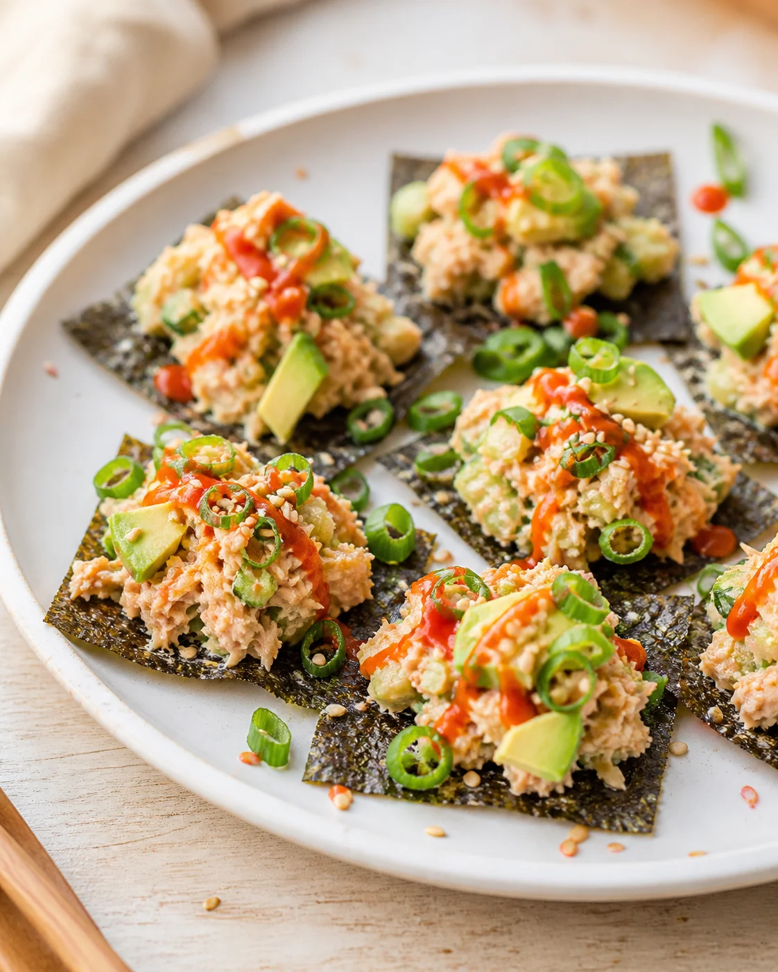 Small crispy nori sheets topped with tuna-avocado mixture, scallions, sesame, and red sauce on a white plate.