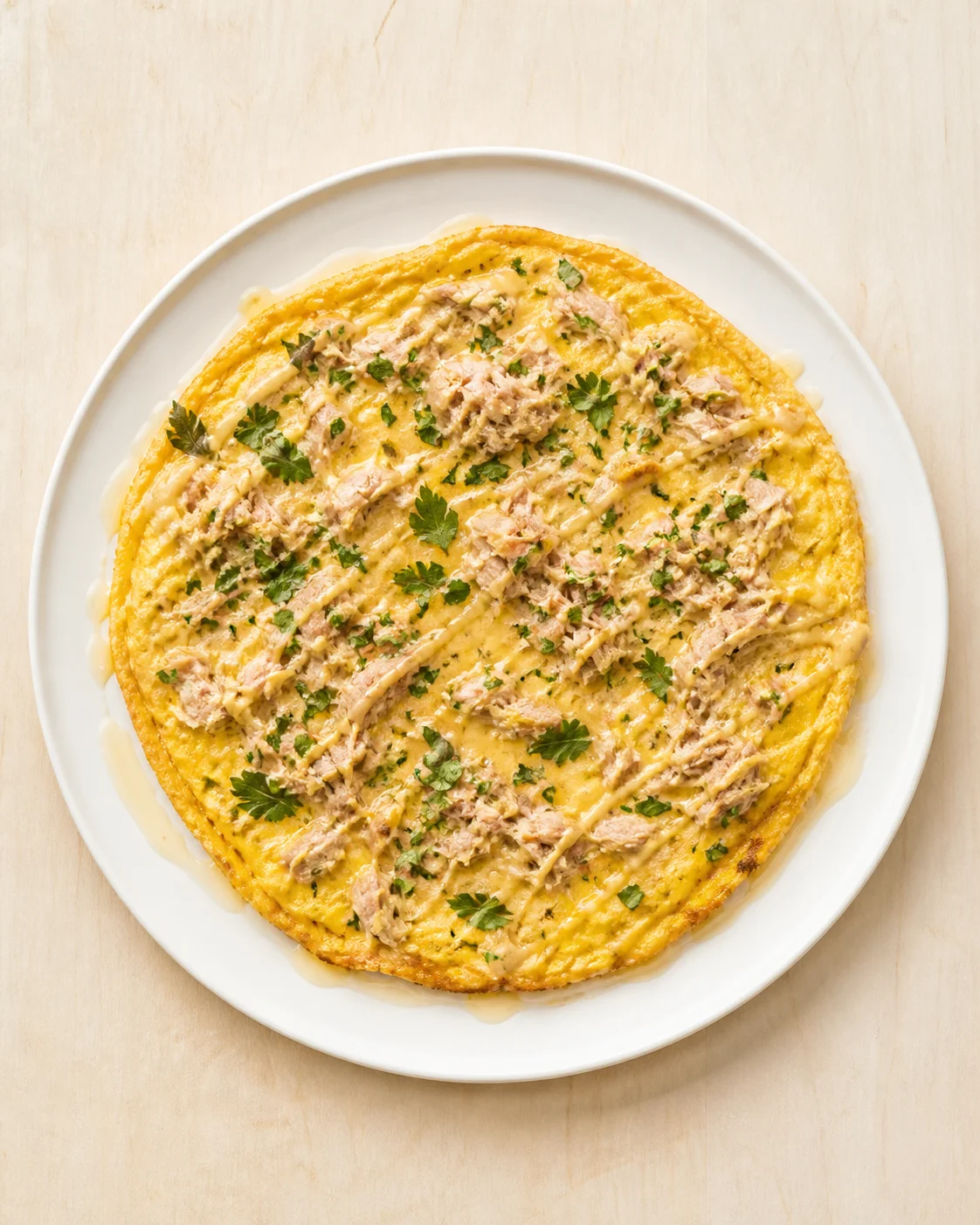 Overhead view of a golden omelette topped with tuna, parsley, and cheese on a white plate.
