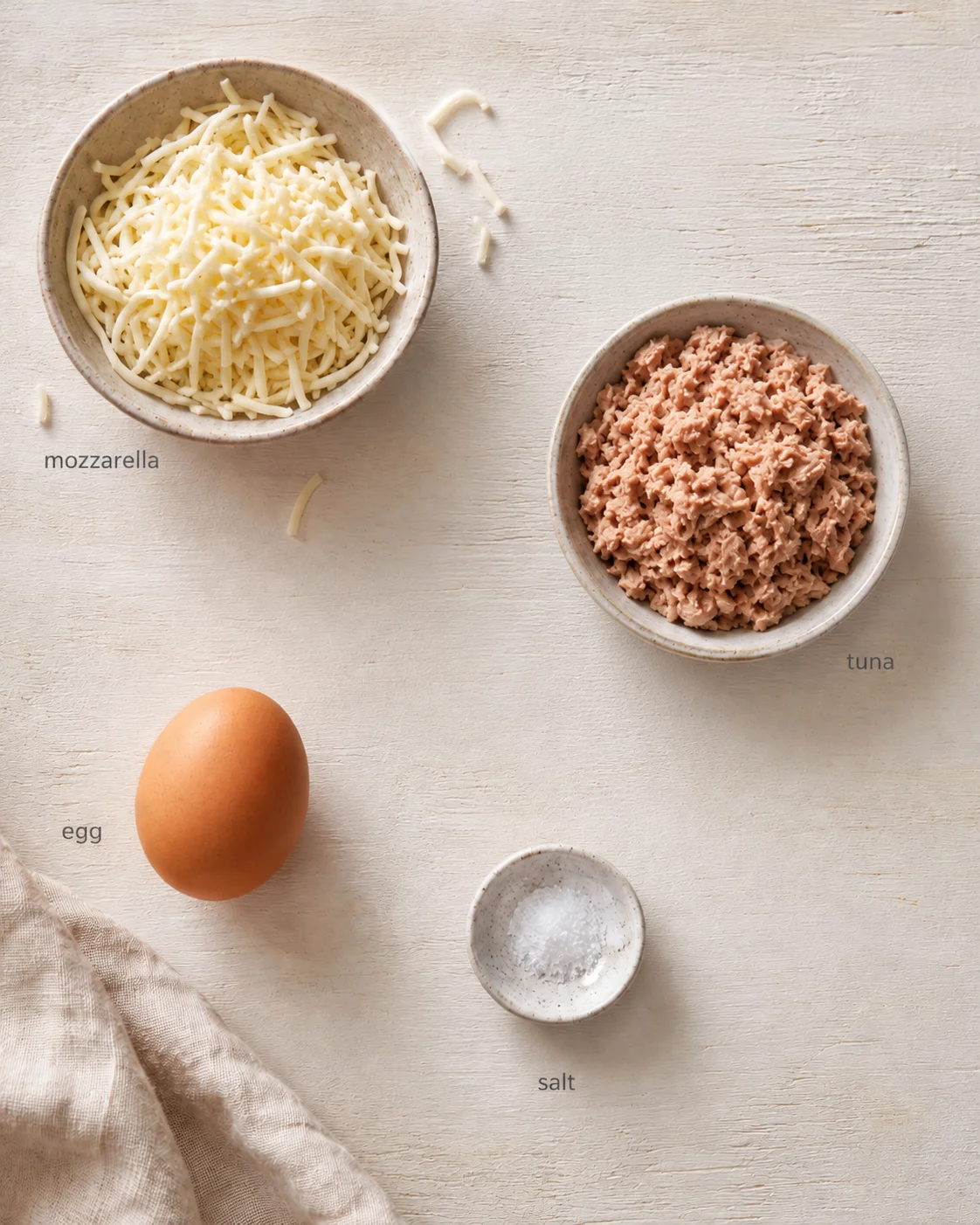 top-down view of shredded mozzarella, tuna, an egg, and a salt dish on a light wooden surface.