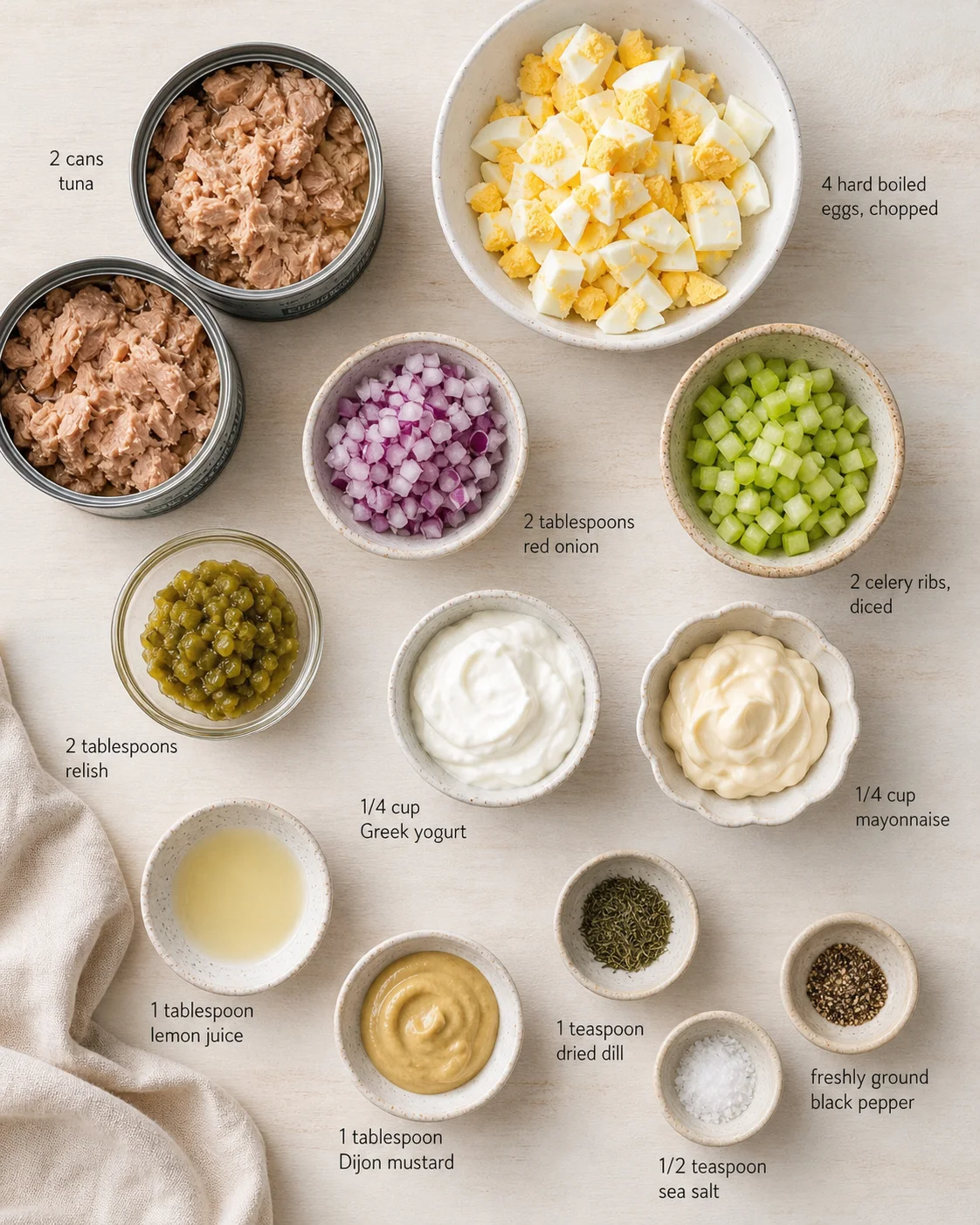 Overhead view of tuna cans, chopped eggs, onions, celery, and condiments arranged in bowls.