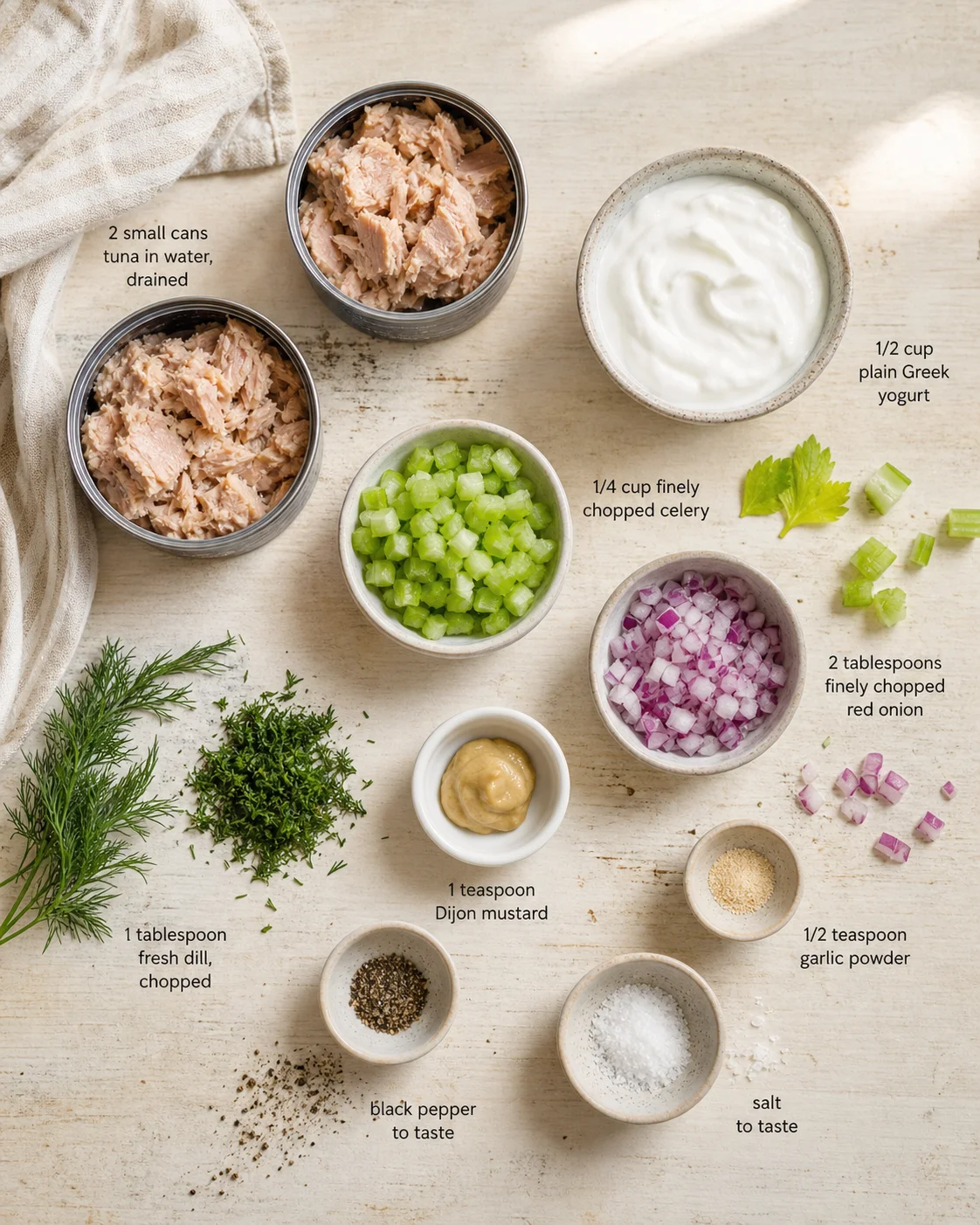 Overhead view of tuna cans, yogurt, celery, red onion, herbs, and spices arranged on a light wooden surface.