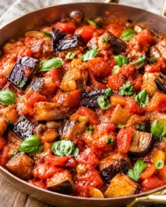 A close-up of a skillet filled with tender eggplant cubes in a rich tomato sauce garnished with basil.