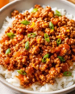 Close-up of a bowl with flavorful orange ground chicken served over rice, garnished with green onions and sesame seeds.