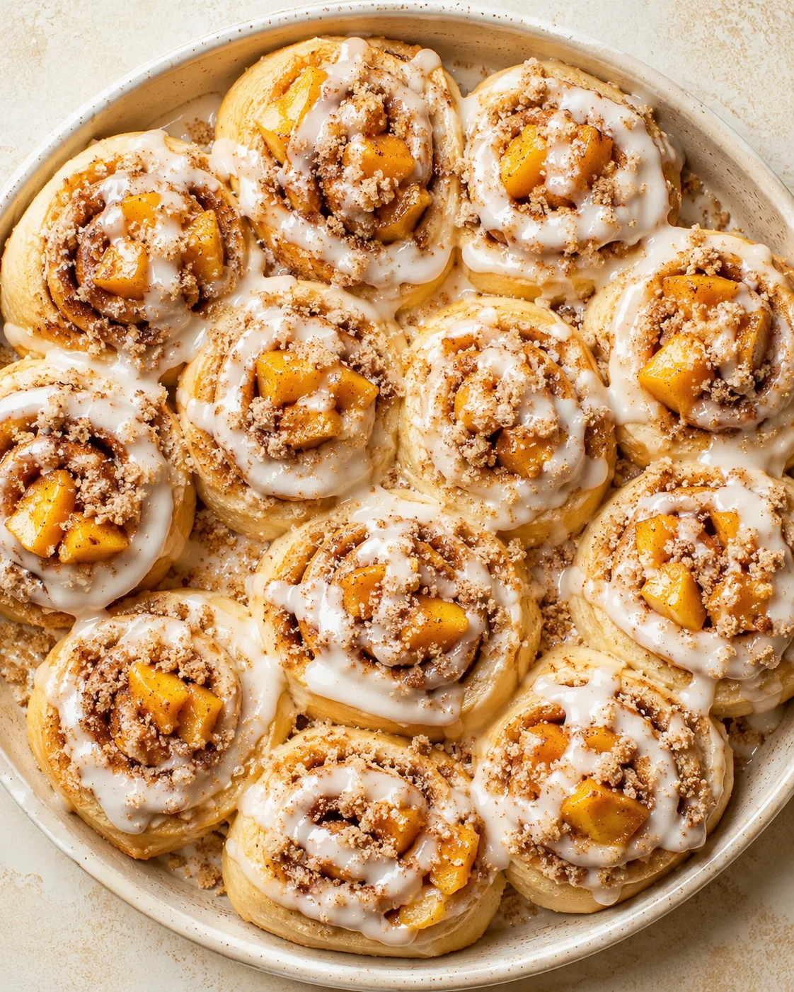 Top-down view of a round baking dish filled with peach-filled cinnamon rolls, white icing, and crumb topping.