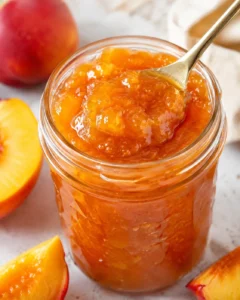 Close-up of vibrant golden nectarine jam in a glass jar with fresh nectarines nearby.