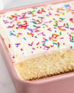 A close-up of a vanilla sheet cake topped with colorful sprinkles and creamy frosting.