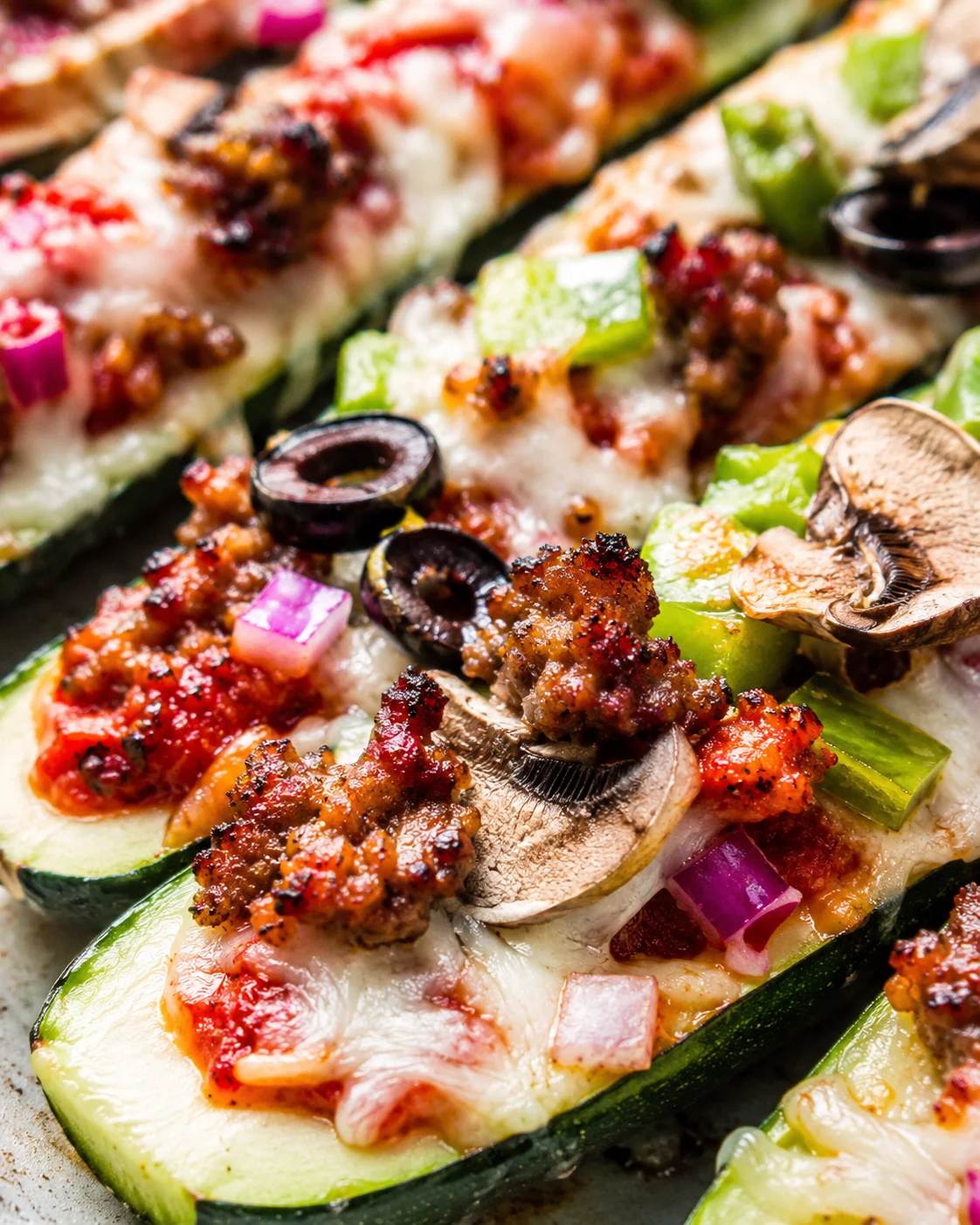 Close-up of cheesy zucchini pizza boats topped with vegetables and olives.