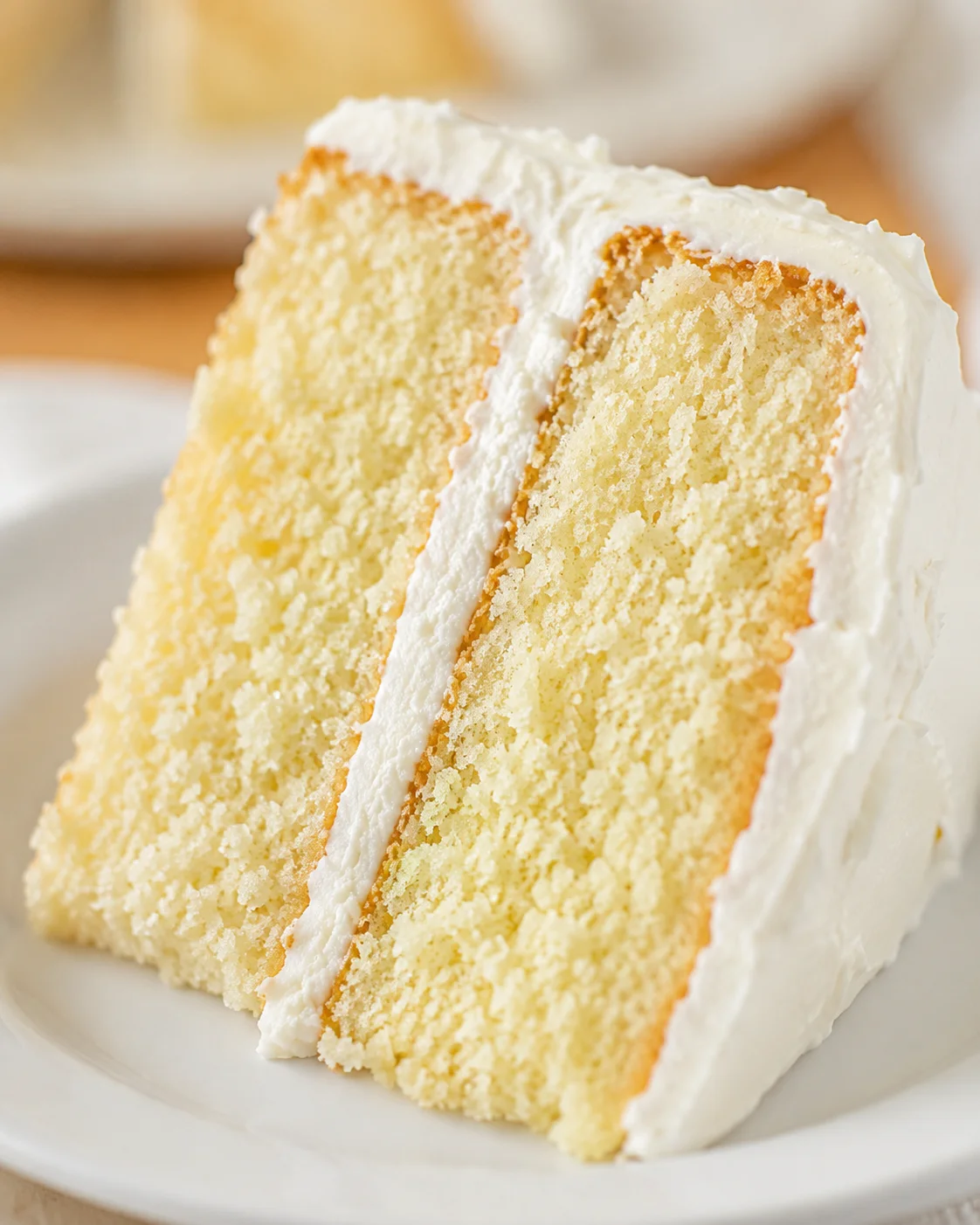 A close-up of a moist vanilla cake slice with creamy frosting on a white plate.