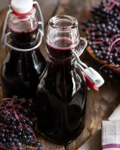 Two swing-top bottles of homemade elderberry syrup with elderberries on a wooden surface