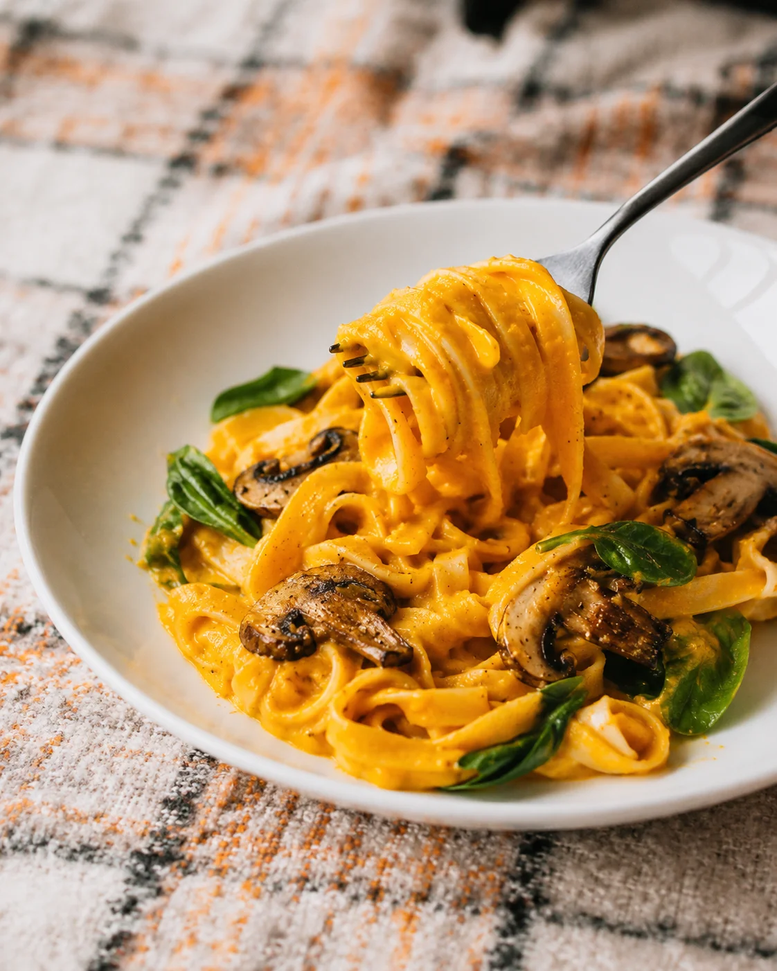 Creamy vegan pumpkin pasta with mushrooms and greens on a white plate.
