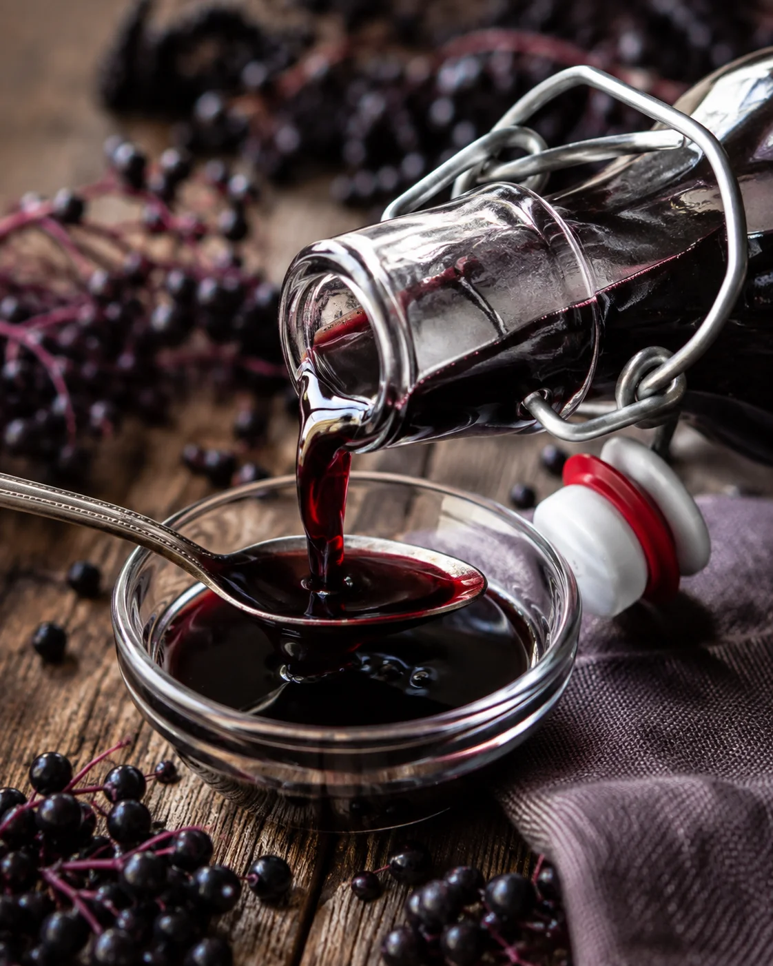 Elderberry syrup being poured from a bottle into a spoon over a small glass bowl with elderberries nearby